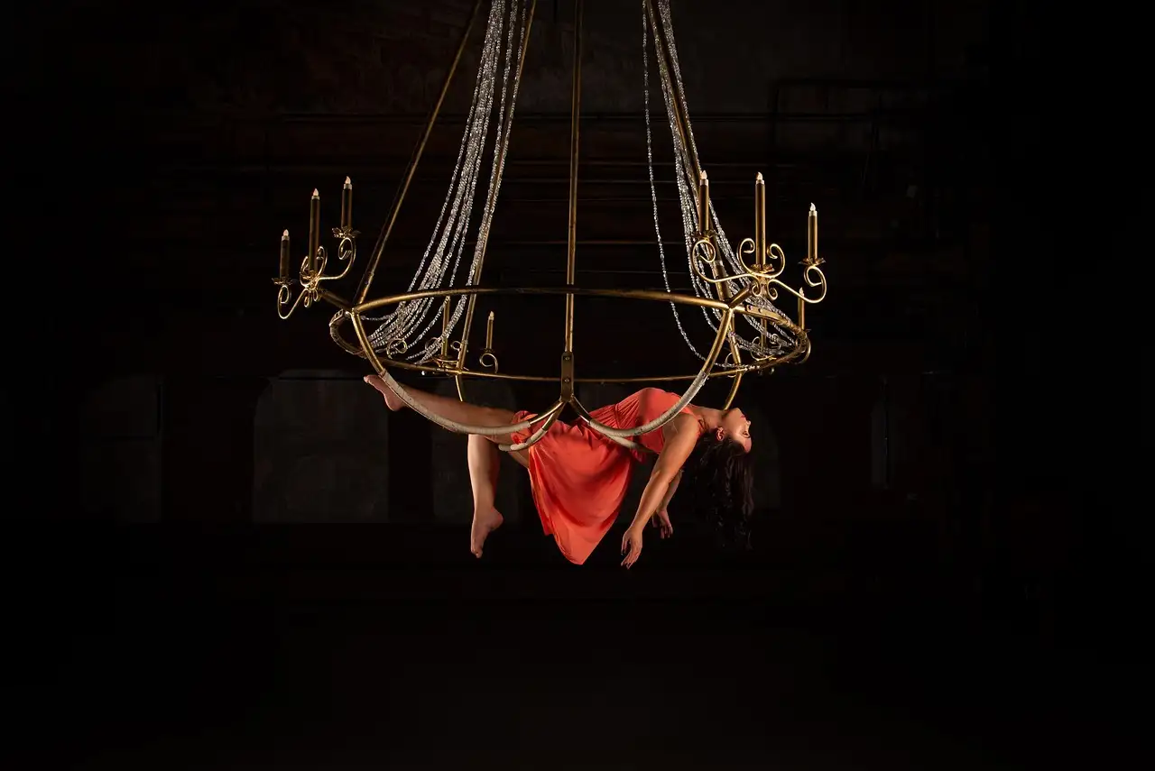 An aerial artist in a coral dress hanging from a gold chandelier hoop under a spotlight in the dark