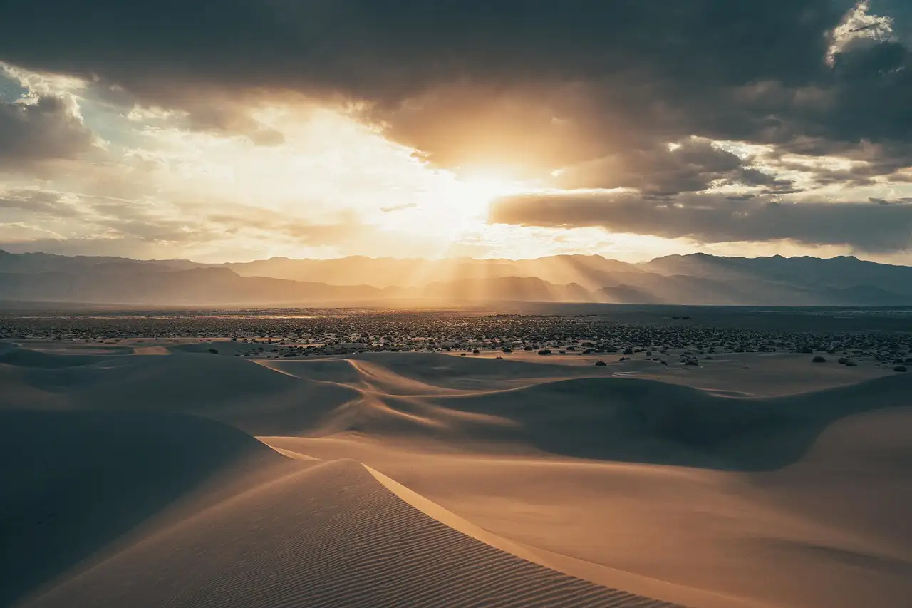 Sand dunes in low evening light under a cloudy sky with rays of sun