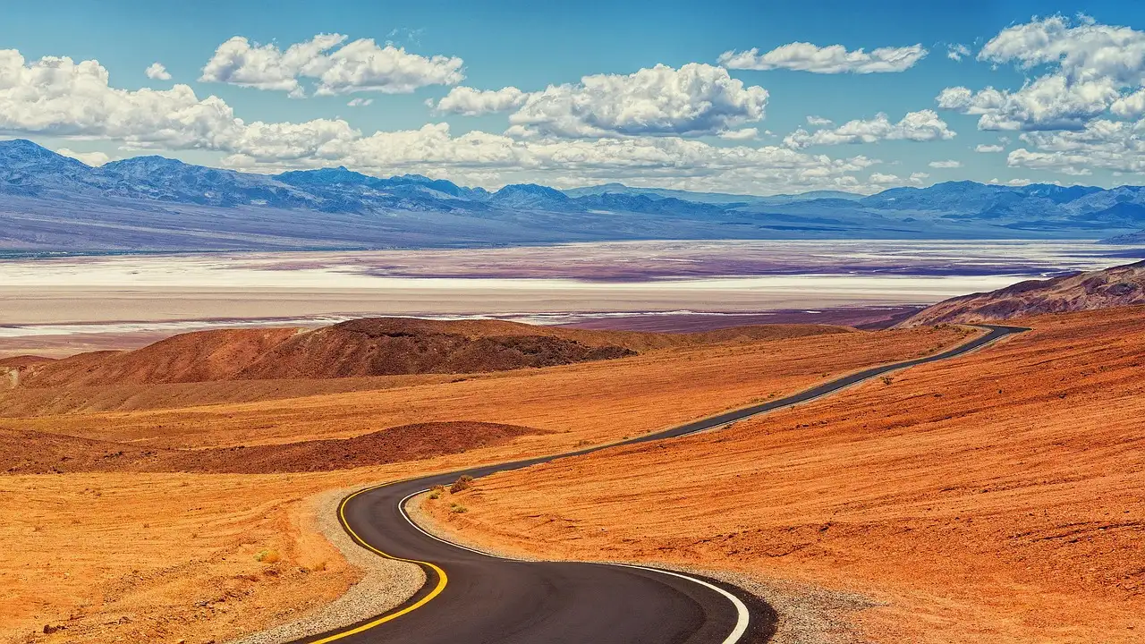 A road curving through orange desert hills toward a white salt flat and distant mountains