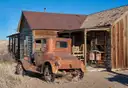 A rusted old car in front of a weathered wooden cabin in a dry desert ghost town