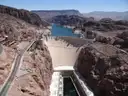 Hoover Dam's curved concrete wall in its canyon, seen from above, with Lake Mead behind