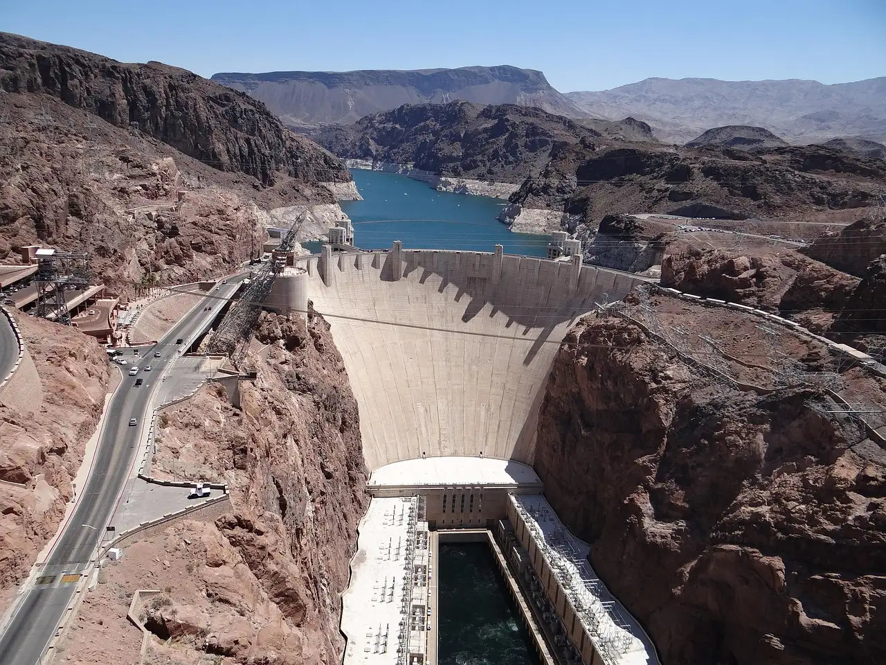 Hoover Dam's curved concrete wall in its canyon, seen from above, with Lake Mead behind