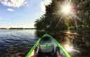 The bow of a green kayak on calm water, seen from the paddler's seat, with trees and sun