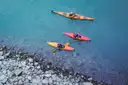 Three bright kayaks seen from above on clear blue-green water next to a rocky shore