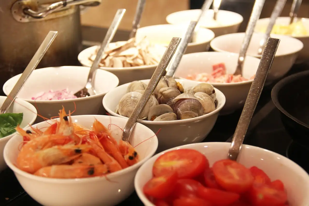White bowls of shrimp, clams, tomatoes and salad with serving spoons on a buffet counter