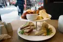 Two-tier afternoon tea stand with finger sandwiches, scones and cake beside a white teapot