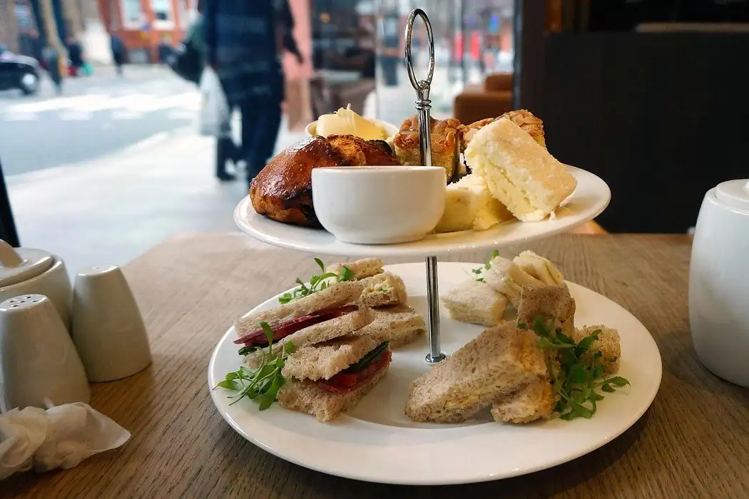 Two-tier afternoon tea stand with finger sandwiches, scones and cake beside a white teapot
