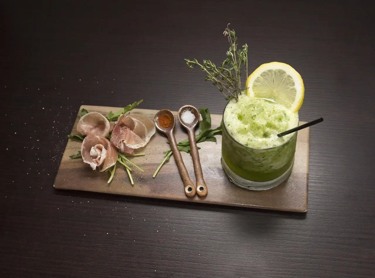 Wooden board with rolled prosciutto and arugula beside a green cocktail garnished with lemon and thyme