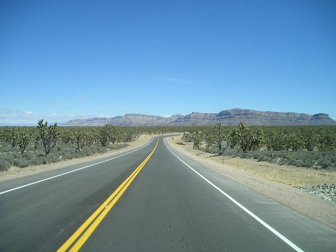 Two-lane desert highway between Joshua trees toward flat-topped cliffs