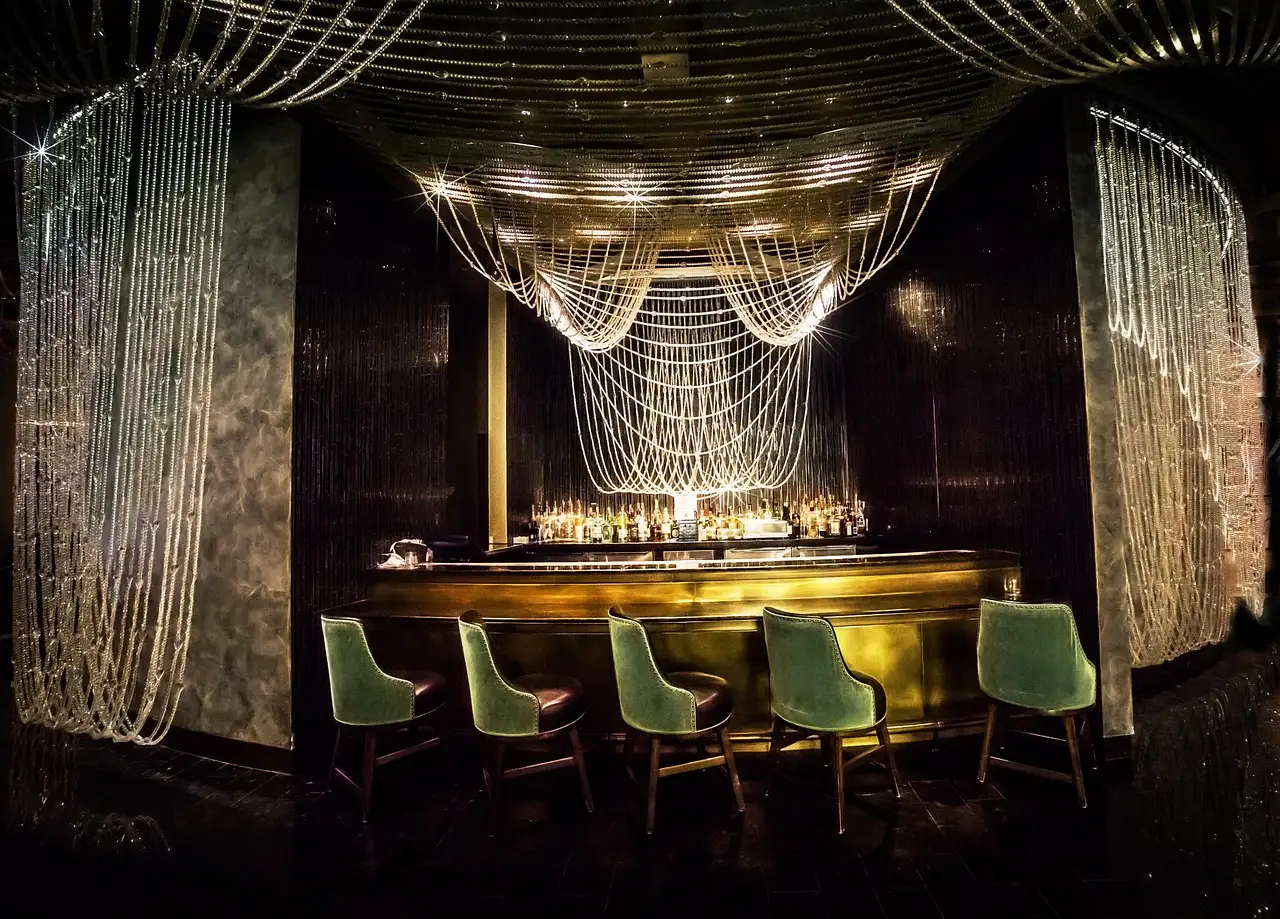 Dim, empty lounge bar with green velvet stools, a glowing gold counter and hanging chain curtains