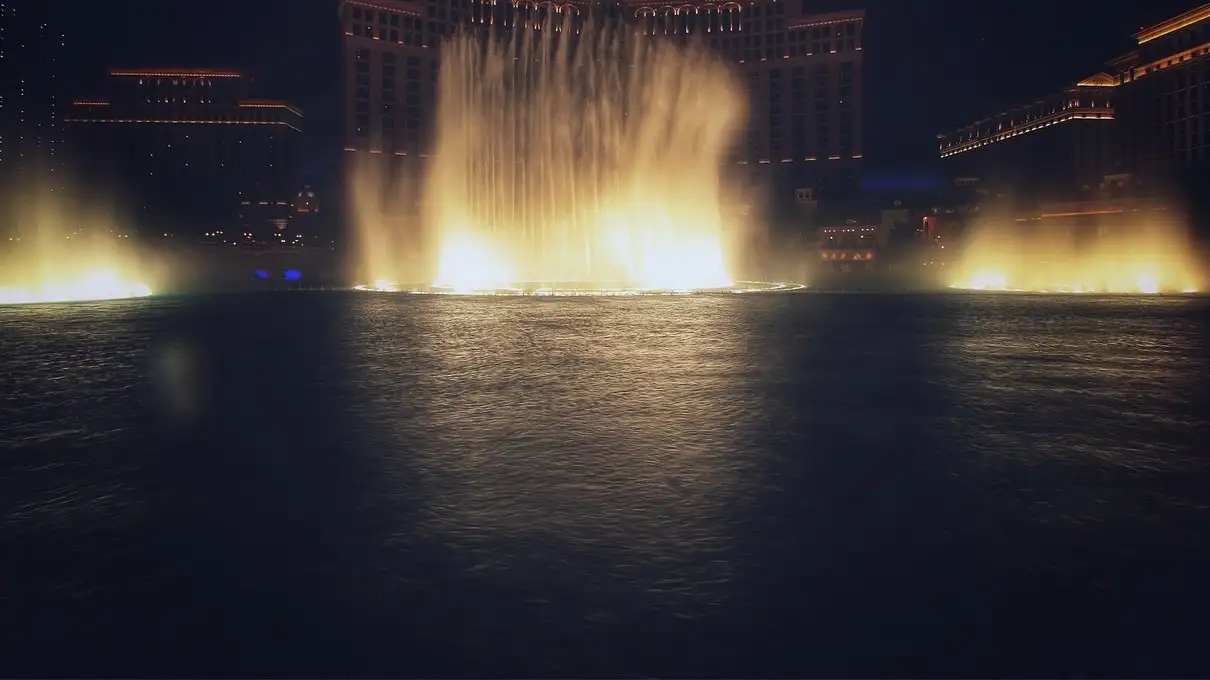 Dancing fountain jets lit gold over a dark lake in front of a resort hotel at night