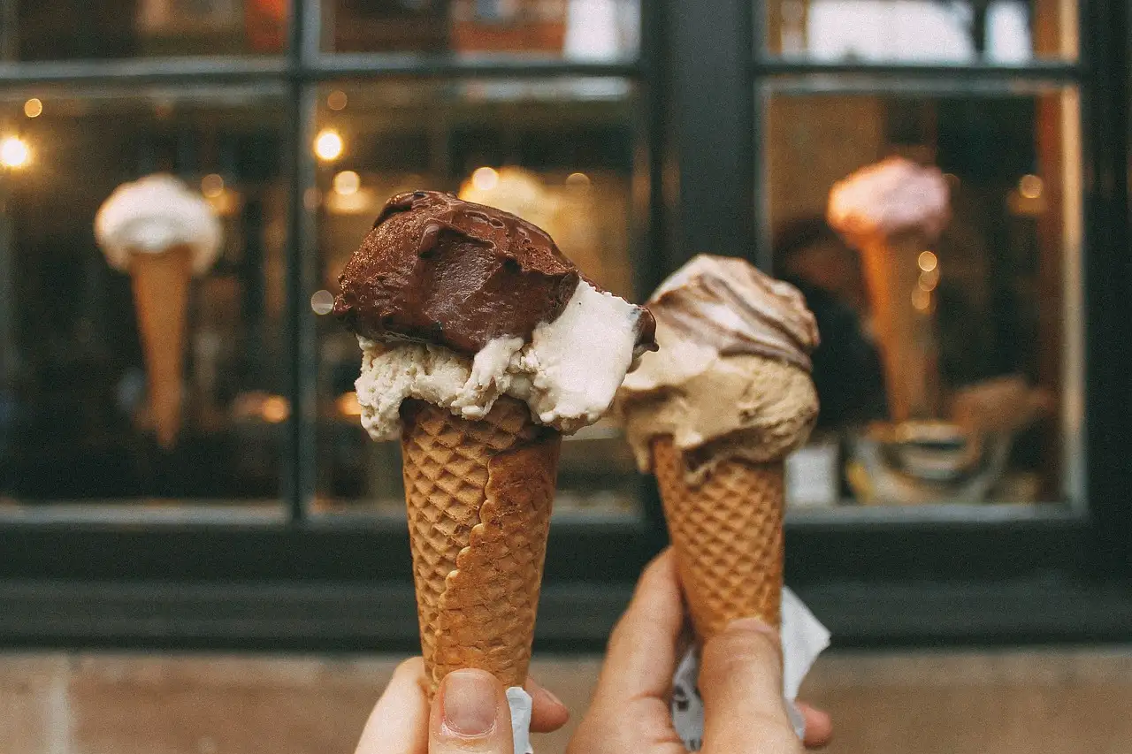 Two hands holding gelato cones in front of a shop window