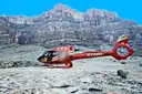 Red helicopter parked on a rocky canyon floor below tall cliffs