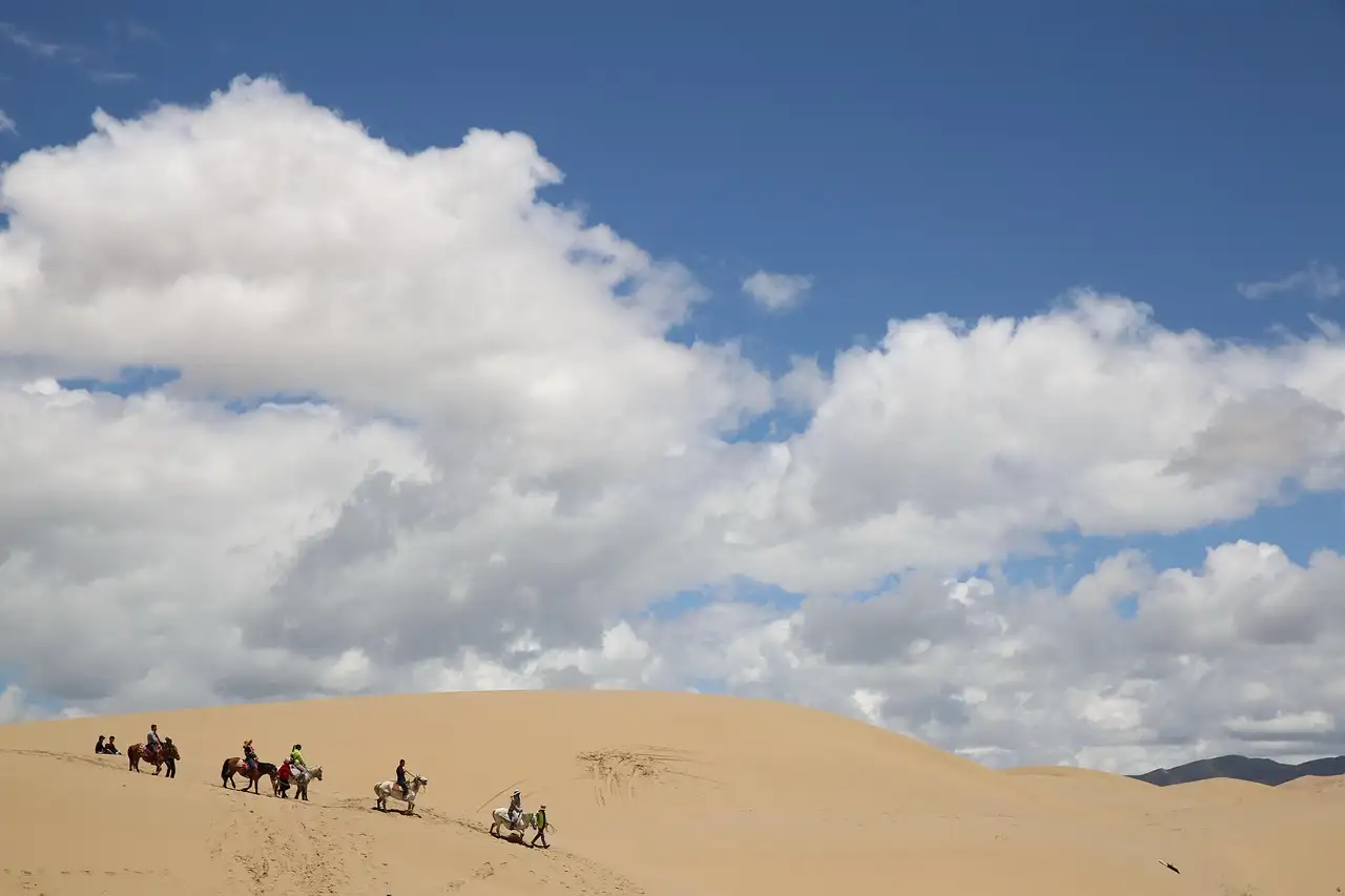 Line of horseback riders crossing pale sand dunes under white clouds