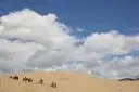 Line of horseback riders crossing pale sand dunes under white clouds