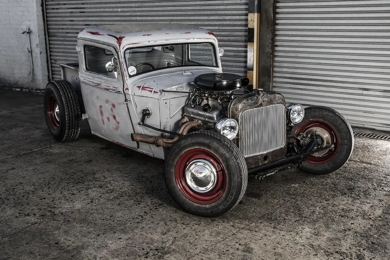 Custom rat-rod pickup with exposed engine and red wheels in front of garage shutters