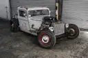 Custom rat-rod pickup with exposed engine and red wheels in front of garage shutters