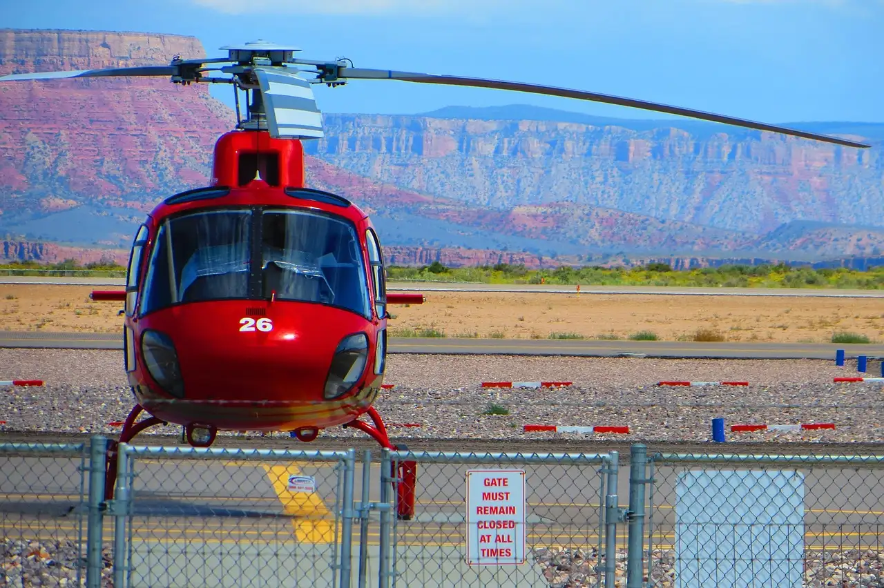 A red tour helicopter parked on a desert helipad behind a fence, with canyon cliffs in the distance