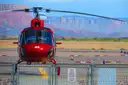 Red helicopter on a desert heliport behind a chain-link gate, cliffs beyond