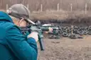 A shooter in a cap firing a carbine at an outdoor range, old tires as a backstop