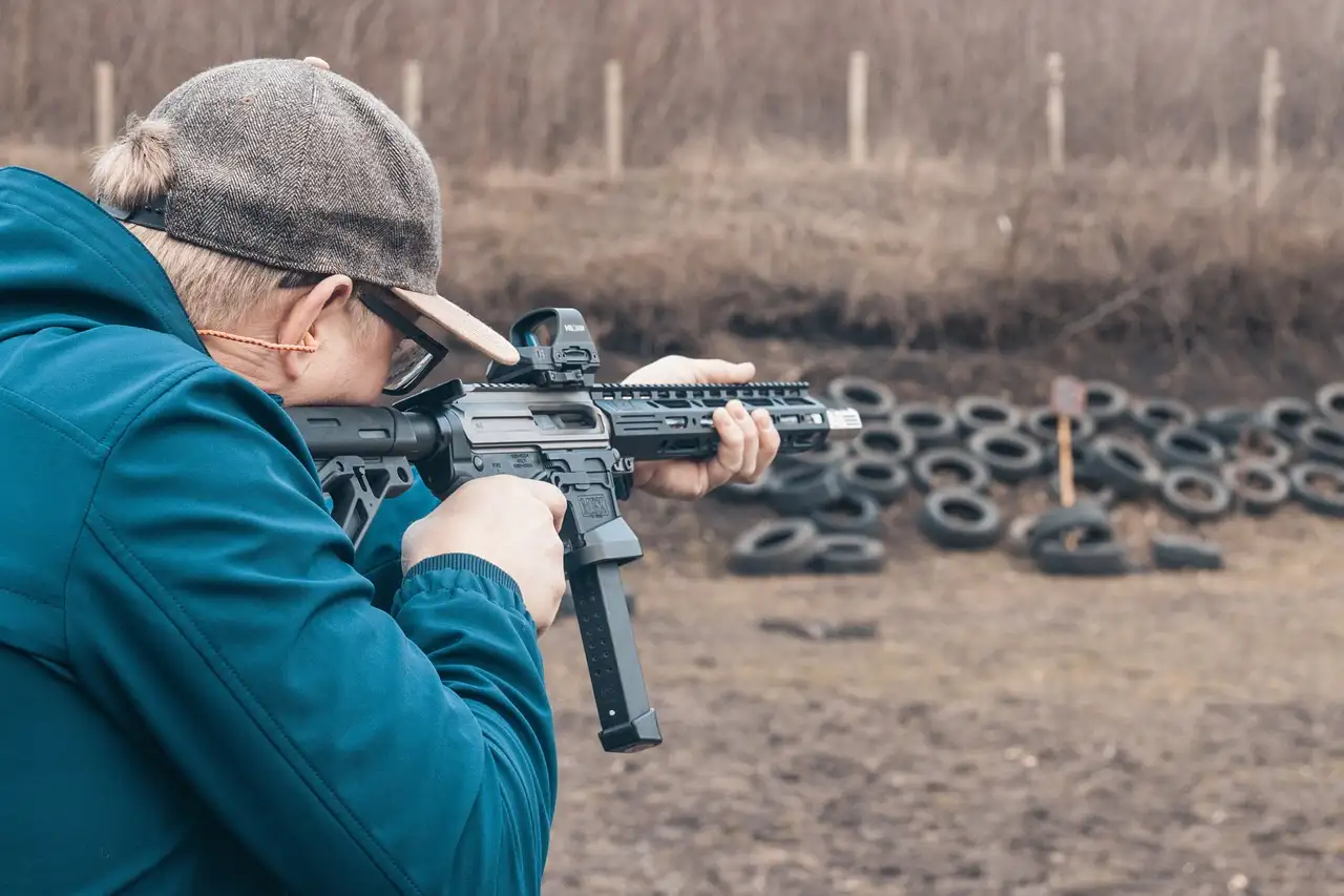 A shooter in a cap firing a carbine at an outdoor range, old tires as a backstop