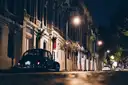 Dark vintage car parked on a lamp-lit city street at night, seen from road level
