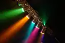 Stage spotlights on a truss shining green, orange, pink and purple beams through haze