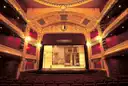 A gilded theatre with a lit stage, red curtains, red seats and stage lights on the balconies