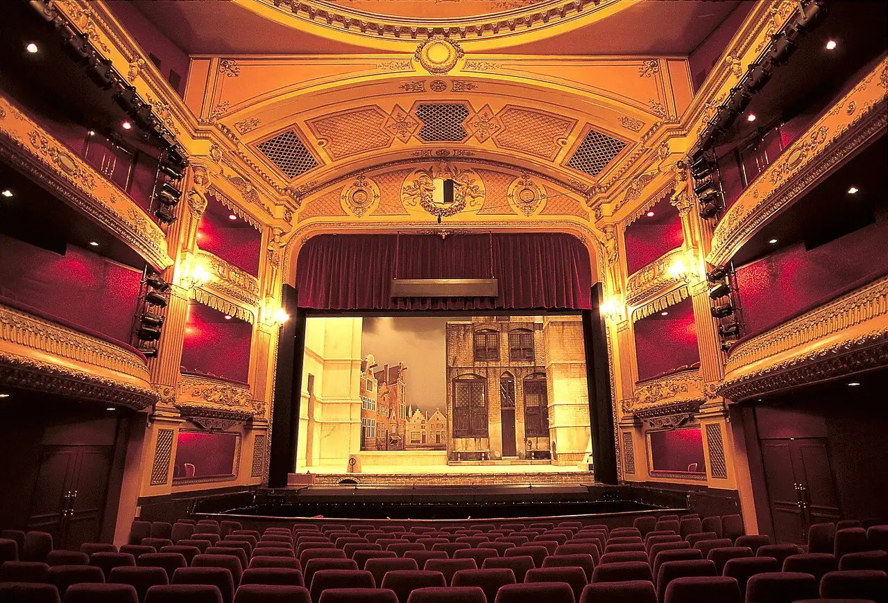A gilded theatre with a lit stage, red curtains, red seats and stage lights on the balconies